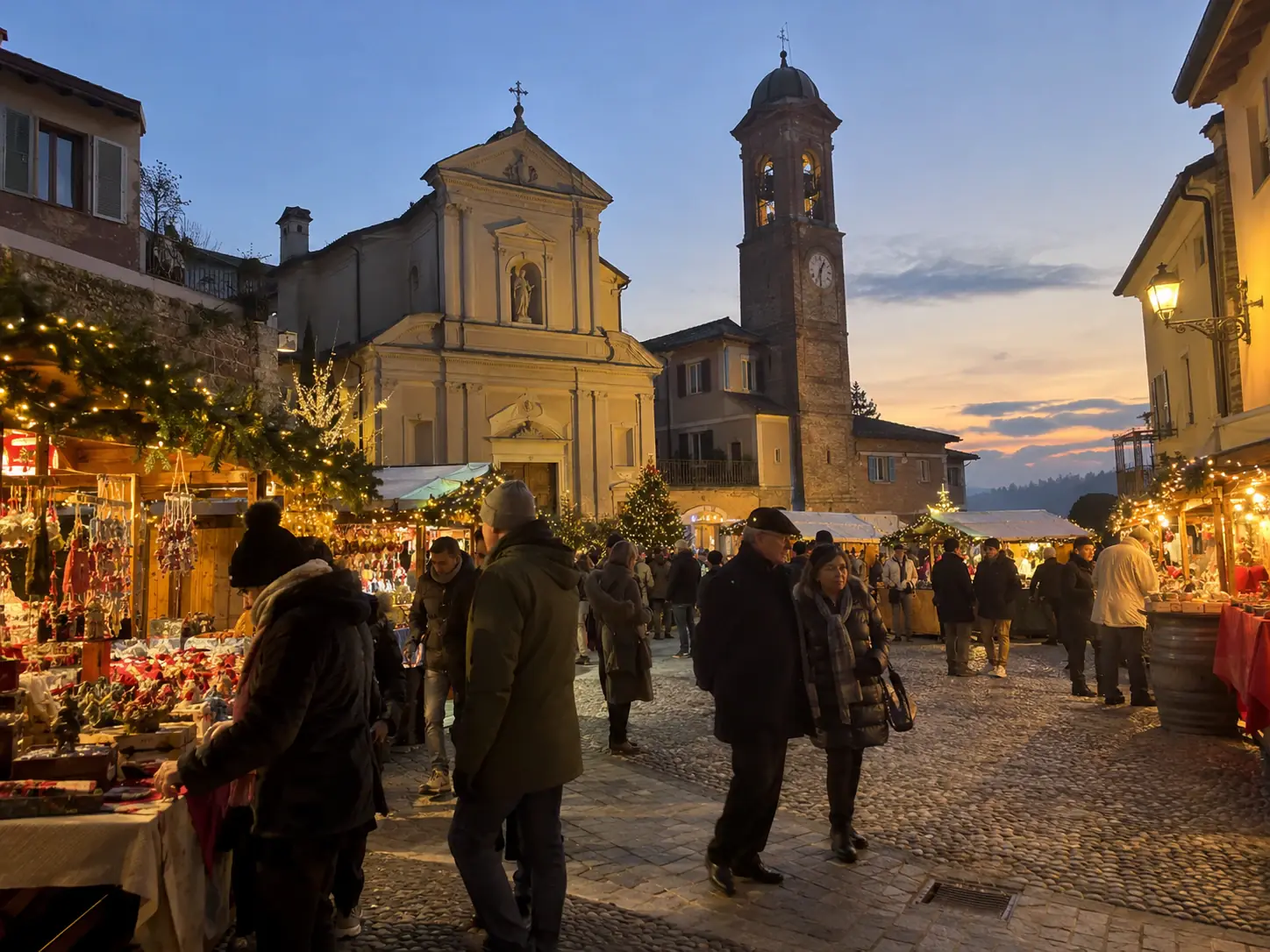 Bancarelle di Natale in una piazza di borgo con chiesa e campanile in mattoni al tramonto, mercatini di Castelletto Monferrato