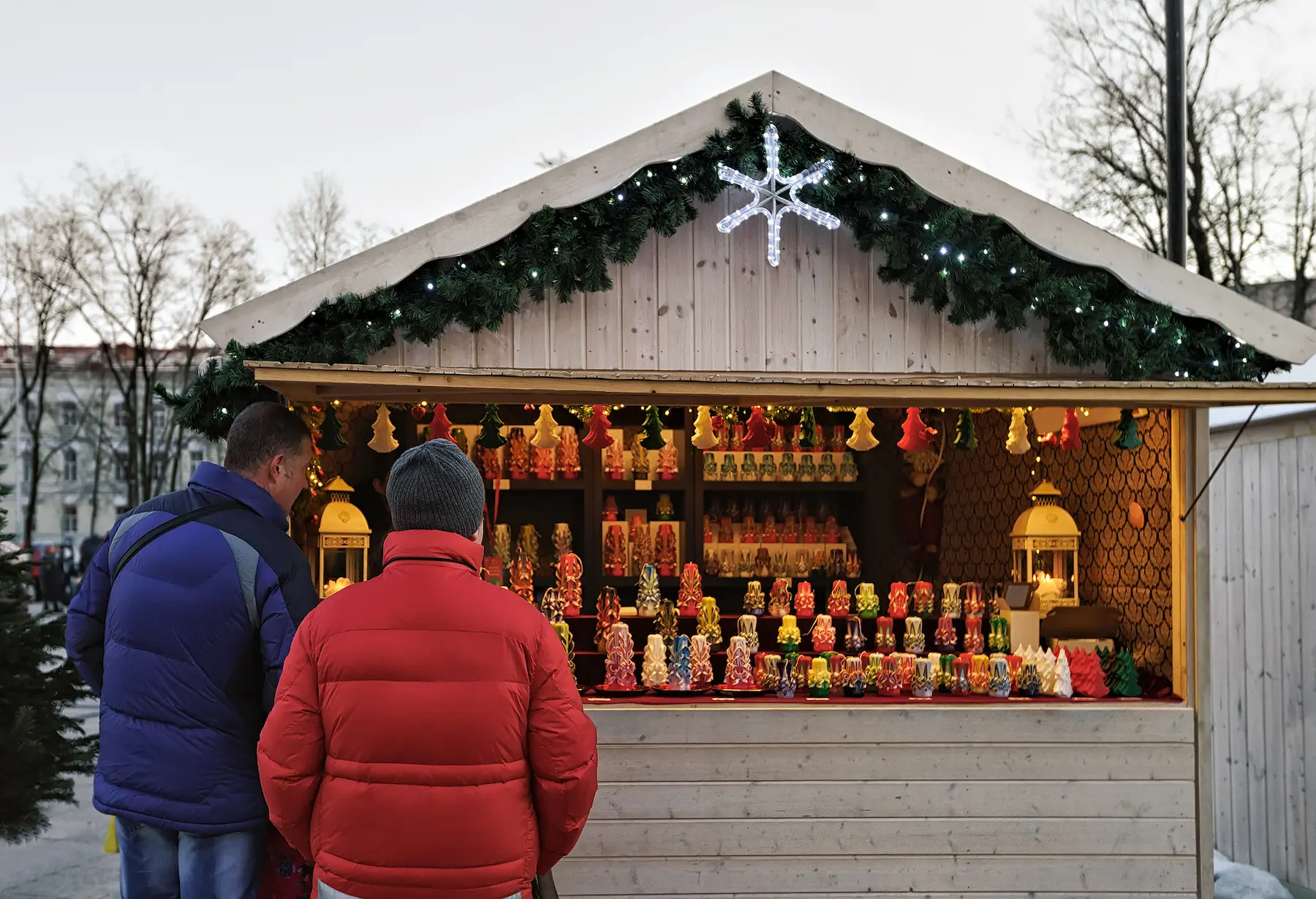 Casetta di legno con ghirlanda e fiocco di neve luminoso, con due visitatori che guardano i prodotti esposti - mercatini di Natale di Cesenatico