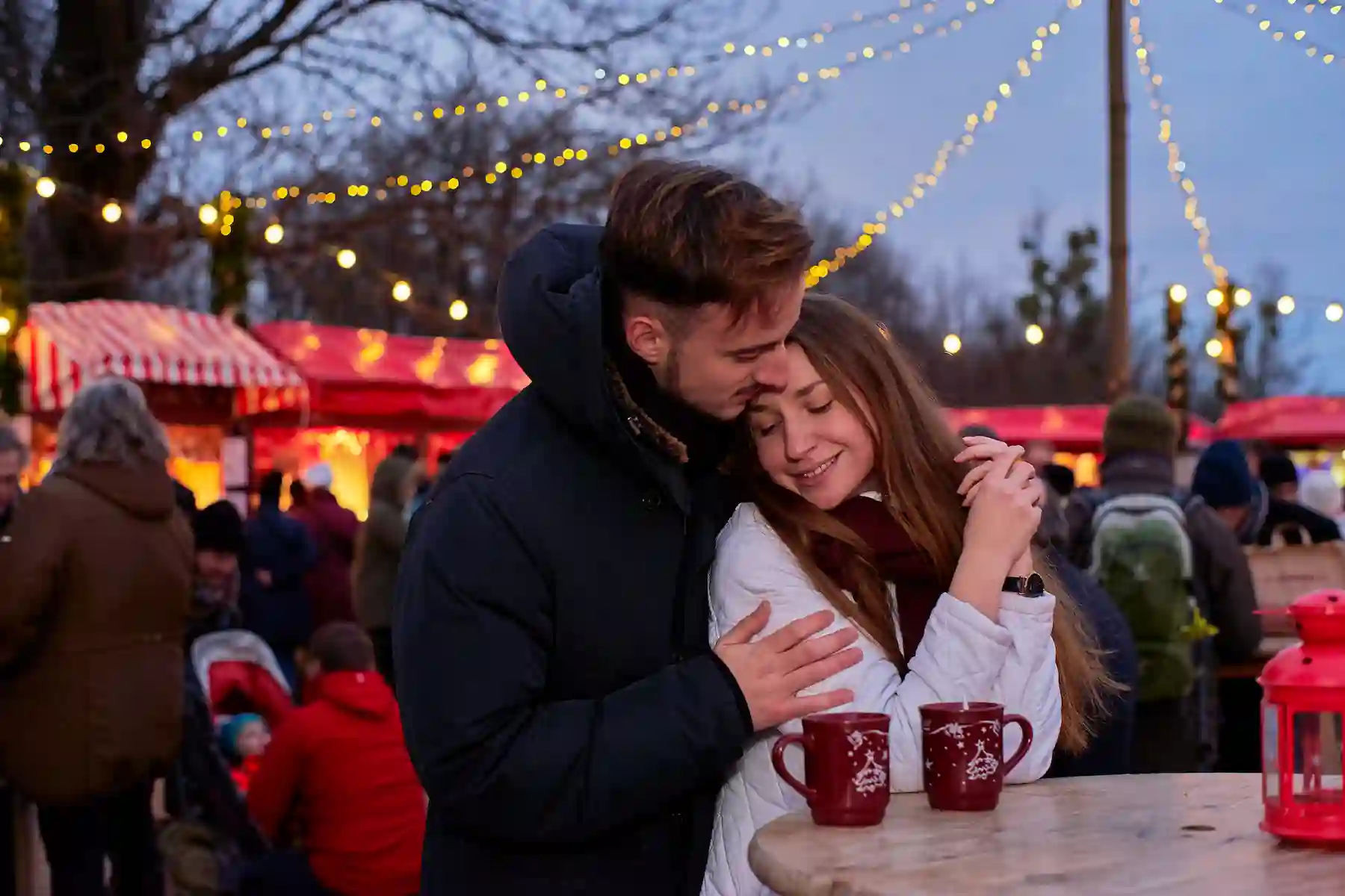 Coppia abbracciata con tazze rosse calde davanti alle casette illuminate - mercatini di Natale di Hône
