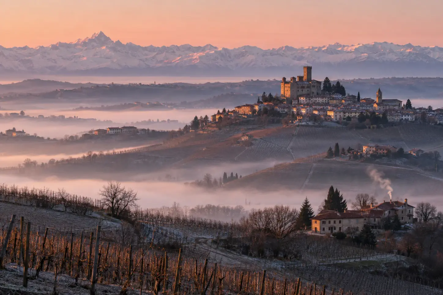 Le colline delle Langhe in inverno con i filari spogli, un borgo con il castello sulla cima, la nebbia nelle valli e le Alpi innevate all'orizzonte